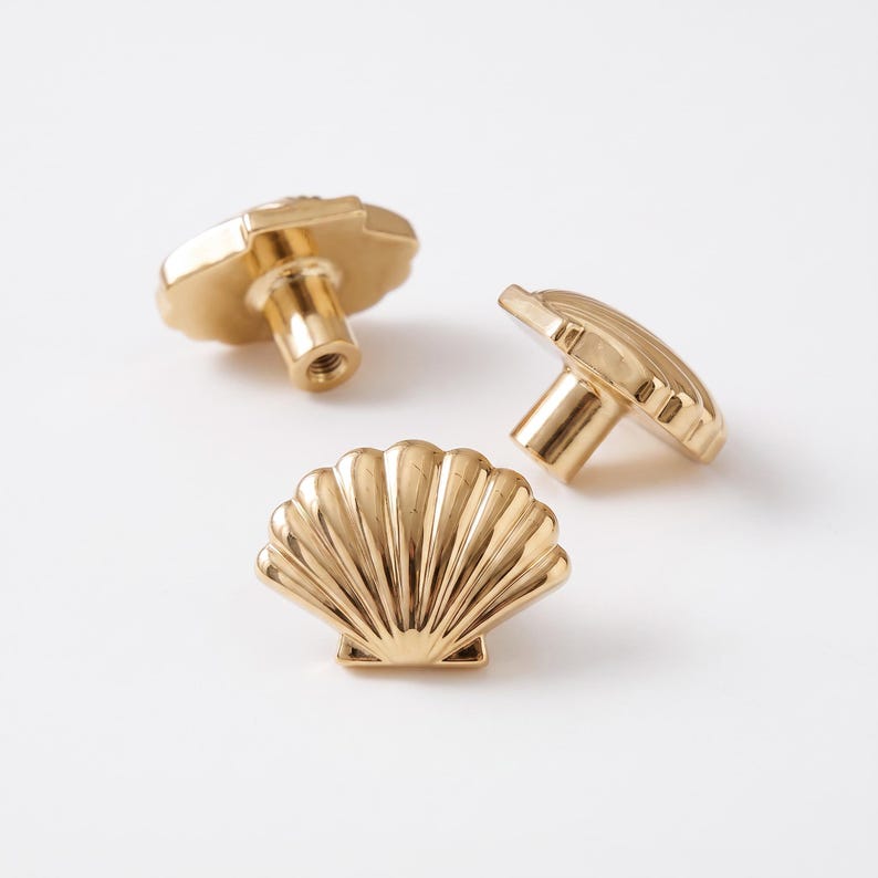 May include: Three gold-colored metal cabinet knobs shaped like seashells. Each knob features a fan-like design with vertical ridges and a cylindrical base with a threaded hole for mounting. The finish is smooth, reflective, and metallic gold.