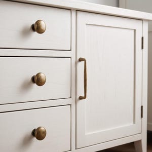 May include: A white wooden cabinet featuring three stacked drawers on the left side and a single door on the right. The drawers are equipped with round, antique brass-toned knobs, while the door features a vertical, matching brass-toned handle.