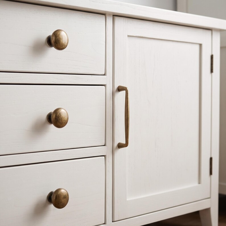 May include: A white wooden cabinet featuring three stacked drawers on the left side and a single door on the right. The drawers are equipped with round, antique brass-toned knobs, while the door features a vertical, matching brass-toned handle.