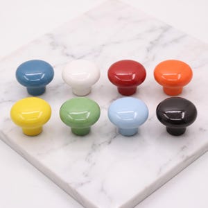 May include: A set of eight round cabinet knobs in a glossy finish, featuring a variety of solid colors including blue, white, red, orange, yellow, green, light blue, and black. Each knob has a smooth, domed top and a cylindrical base.