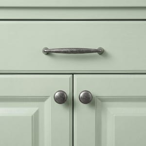 May include: A light sage green cabinet featuring one horizontal drawer with a dark, weathered metal pull handle and two lower cabinet doors, each equipped with a matching dark, weathered metal round knob for opening and closing the doors.