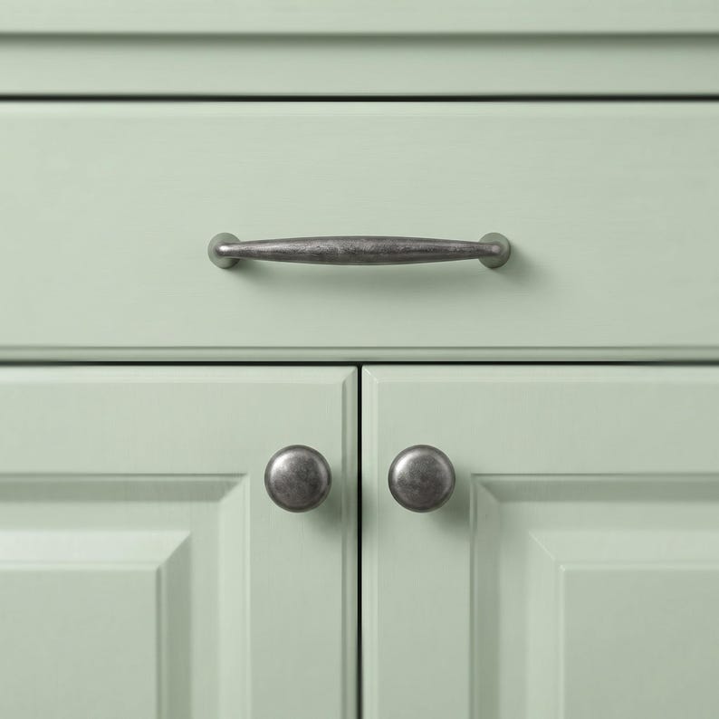 May include: A light sage green cabinet featuring one horizontal drawer with a dark, weathered metal pull handle and two lower cabinet doors, each equipped with a matching dark, weathered metal round knob for opening and closing the doors.