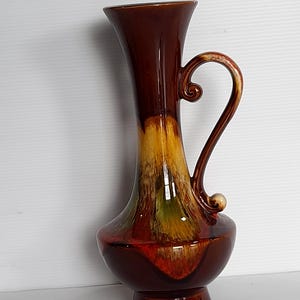 May include: A tall, brown ceramic vase with a handle. The vase has a flared top and a bulbous base. The colors transition from brown to yellow, green, and red. The handle has a decorative swirl.