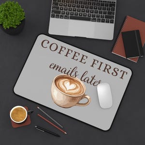 May include: A rectangular desk mat with a light gray surface featuring the brown text "COFFEE FIRST emails later" and a graphic illustration of a ceramic coffee cup filled with latte art in the shape of a heart centered on the mat.