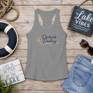 May include: A grey racerback tank top featuring a gold anchor graphic and the dark blue text "Dockside Darling" printed on the front. The design includes a decorative gold swirl accenting the anchor and the text in a script font.