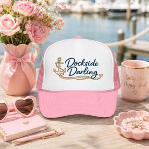May include: A pink and white trucker hat featuring a white front panel with a gold anchor graphic and the dark blue text Dockside Darling. The hat has a solid pink brim and a mesh back, designed with a structured crown and an adjustable snapback closure.