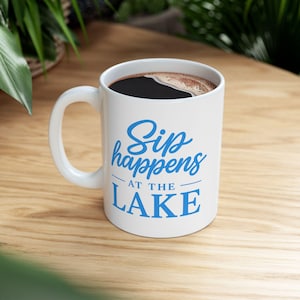 May include: White ceramic mug filled with dark coffee, featuring the blue text "Sip happens at the lake." The mug has a white handle and sits on a light wooden surface. The background includes green foliage.