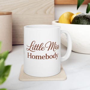 May include: A white ceramic mug with the words "Little Miss Homebody" in brown script. The mug is on a beige coaster, with a blurred background including a wooden surface and a bowl of fruit.