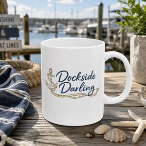May include: A white ceramic mug featuring a navy blue and gold graphic of an anchor with the text Dockside Darling in a script font. The design includes a small gold heart at the end of the text, centered on the front of the mug.
