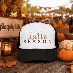 May include: A white and black trucker hat featuring the text "Latte SEASON" in brown lettering on the front panel. The hat has a solid black brim and a white front panel with black mesh sides and back, designed with a structured crown and adjustable strap.