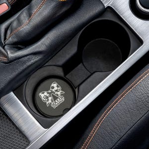 May include: Black car cup holder with a coaster featuring a soccer ball bow design and the text "Soccer Mama" in white. The coaster is placed in the cup holder, which is surrounded by black leather and silver trim.