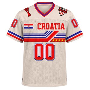 May include: A cream-colored V-neck jersey featuring the text CROATIA in red, the Croatian flag, and the number 00 in red with blue outlines. The design includes red and blue horizontal stripes, five yellow stars, and soccer-themed graphics.