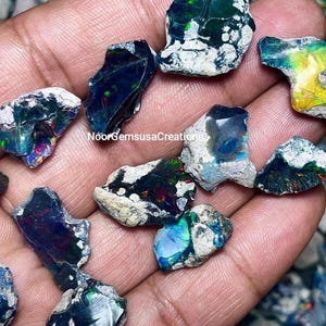 May include: A collection of raw, uncut opals in various shapes and sizes, showcasing a spectrum of colors including blues, greens, and reds. The opals are held in a hand, with the text "NoorGemsusaCreations" visible.