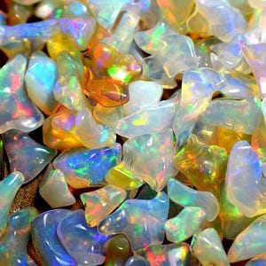 Może przedstawiać: Zbliżenie na kolekcję opalów. Opale mają różne kształty i rozmiary, z półprzezroczystą, mleczną podstawą. Wykazują żywą grę barw, z błyskami niebieskiego, zielonego, żółtego i pomarańczowego. Kamienie szlachetne są rozrzucone.