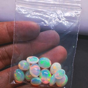 May include: A collection of oval-shaped opals in a clear plastic bag. The opals display a range of colors, including white, orange, green, and blue, with iridescent flashes. The bag is held by a person.