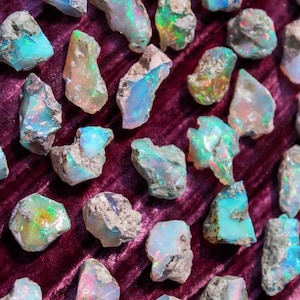 May include: A collection of raw opals with vibrant colors, including blues, greens, and oranges, displayed on a deep purple velvet surface. The opals have a rough, natural texture and varying shapes, showcasing their unique iridescence.