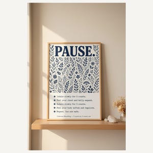 May include: A rectangular wall art print in a light wood frame featuring the word PAUSE in large dark blue letters above a detailed botanical illustration of leaves and flowers. Below the illustration are five breathing instructions in dark blue text.