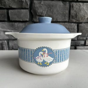 Pode incluir: Uma caçarola de cerâmica branca com tampa azul-clara a condizer e pegas laterais. O exterior apresenta um medalhão circular central que retrata dois gansos brancos com fitas cor-de-rosa, rodeado por um padrão de borda com riscas azuis.