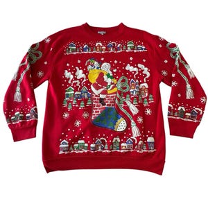Puede incluir: Una sudadera roja de manga larga con un gráfico de Santa Claus sentado sobre una chimenea de ladrillo con un saco de regalos. El diseño incluye un calcetín azul, casas de pueblo nevadas, copos de nieve blancos y cordones verdes con borlas.