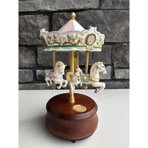 May include: A decorative carousel music box featuring two white horses with pastel saddles and floral accents. The canopy is painted in soft pink, blue, and white with gold trim, supported by a central gold pole on a round, polished dark wood base.