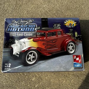 以下が含まれることがあります： 1:25スケールのボイド・コディントン・アメリカン・ホットロン1932フォード・クーペのモデルキット箱。箱には、炎のグラフィック、クロームホイール、AMT ERTLロゴが描かれた赤と白の車が写っています。テキストにはSkill 2、10+、モデルキットの詳細が含まれています。