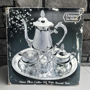 May include: A silver-toned coffee set on a round tray, featuring a coffee pot, a creamer, and a sugar bowl with lids. The packaging displays the text Elegance In Silver and Three Piece Coffee Set With Round Tray against a dark background.