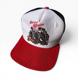 Könnte beinhalten: Eine weiß-schwarze Trucker-Cap mit rotem Schirm. Auf der Vorderseite befindet sich eine Grafik eines Monstertrucks, der kleinere Fahrzeuge überrollt, sowie der rote Schriftzug "...there is a difference" und das "Snap-on"-Logo.