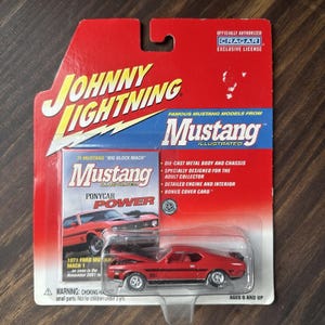 May include: A red 1971 Ford Mustang Mach 1 die-cast model car in its original packaging. The packaging features the Johnny Lightning logo, Mustang Illustrated branding, and details about the model's features.