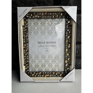 May include: A rectangular picture frame featuring a border encrusted with amber, brown, and clear faceted jewels. The frame is housed in white cardboard packaging labeled Belle Maison Splendid Jewel Collection 5x7. The insert displays a repeating pattern.