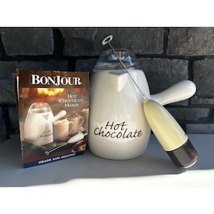 Puede incluir: Una chocolatera de cerámica blanca con asa y el texto Hot chocolate impreso en marrón, acompañada de un espumador eléctrico de mano con mango color crema y un folleto de instrucciones que muestra la misma chocolatera.