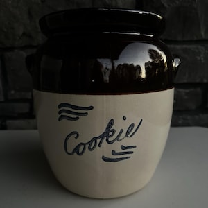 May include: A ceramic cookie jar with a two-tone design featuring a dark brown glazed upper section and a cream-colored lower body. The word Cookie is inscribed in dark blue script on the front, accompanied by decorative wavy lines.