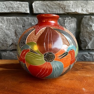 May include: A round ceramic vase featuring a smooth, polished finish with hand-painted designs of hummingbirds and large flowers in shades of red, green, blue, and brown, set against a textured, tan-colored background.