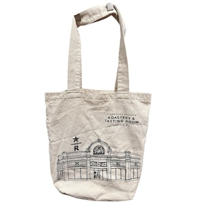 May include: Beige canvas tote bag with black graphic of a building and text that reads "STARBUCKS RESERVE ROASTERY & TASTING ROOM SEATTLE, WA". The bag has two handles and a small clasp at the top.
