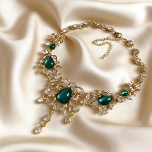 May include: A gold-toned necklace with emerald green teardrop-shaped gemstones and clear crystals. The necklace features floral and leaf-like designs, with a delicate chain closure. The gemstones and crystals are set in a gold metal.