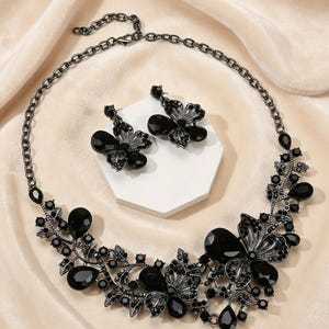 May include: A black floral necklace and matching earrings set. The necklace features large, teardrop-shaped black gemstones and smaller, round black stones. The earrings mirror the floral design of the necklace. The jewelry is set against a neutral background.