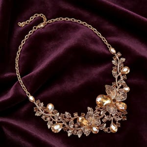 Peut inclure: Collier doré orné de motifs floraux et de feuilles, orné de pierres précieuses de couleur ambre. Le collier est doté d'une fine chaîne et se détache sur un fond violet foncé. Les éléments floraux sont au centre du bijou.