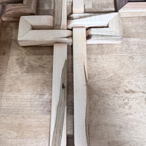 May include: A wooden cross assembled from interlocking pieces. The cross is made of light-colored wood with darker grain patterns. The design features a geometric, angular aesthetic, with a focus on the natural wood grain.