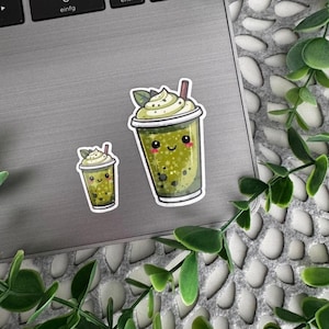 Könnte beinhalten: Zwei gestanzte Aufkleber mit einem grünen Matcha-Bubble-Tea-Getränk, das ein lächelndes Gesicht, rosa Wangen, Schlagsahne und einen Strohhalm hat. Das Design zeigt ein Sternenmuster und dunkle Boba-Perlen in einem durchsichtigen Becher.