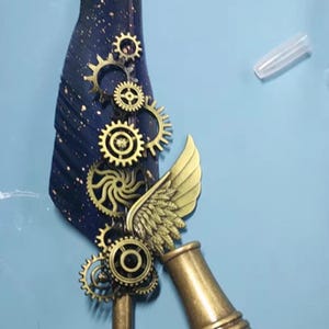 Puede incluir: Un objeto decorativo de estilo steampunk. Presenta un elemento en forma de pluma azul oscuro con motas doradas, engranajes color latón, un ala y una base cilíndrica, todo sobre un fondo azul claro.