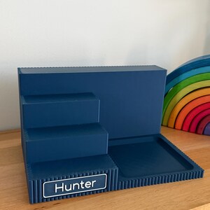 May include: A dark blue tiered display stand featuring three levels and a flat square tray section. The front edge of the bottom tier includes a rectangular nameplate with the white text Hunter printed on a dark blue background with a white border.