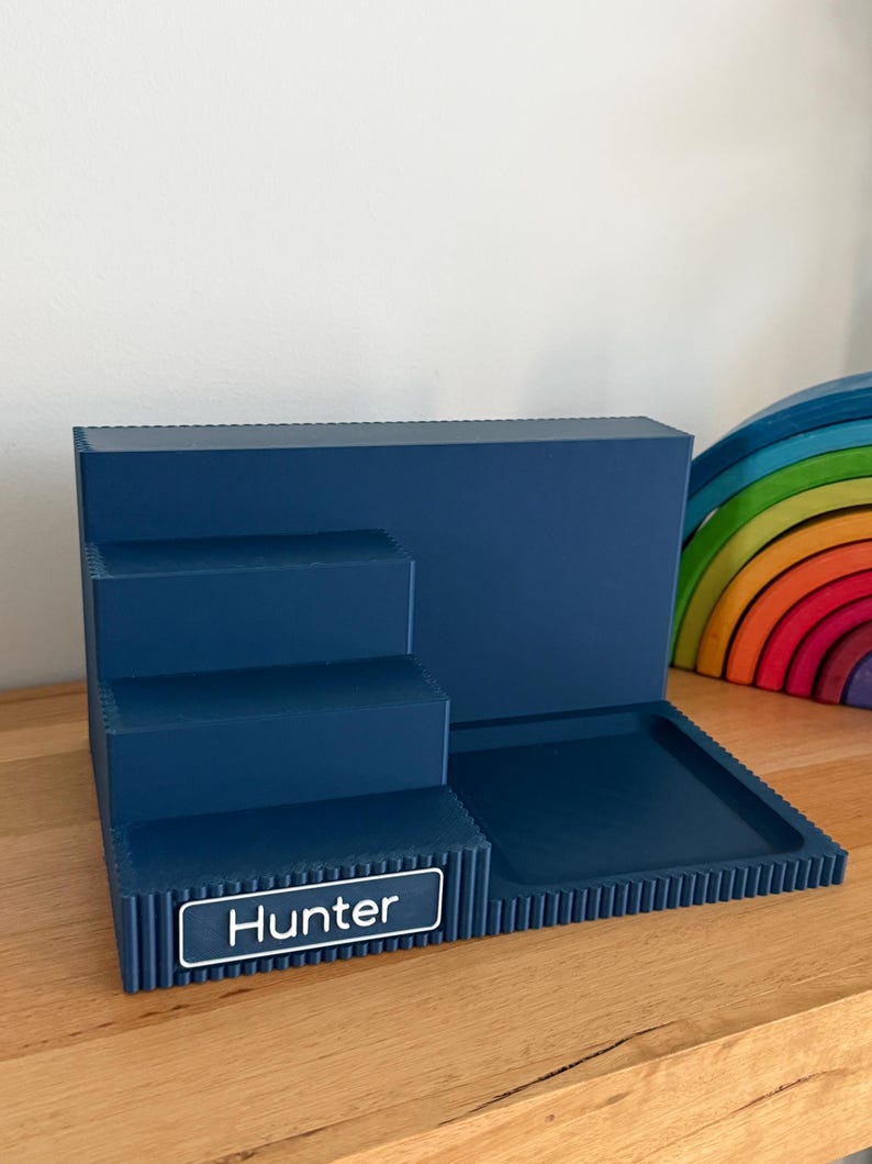 May include: A dark blue tiered display stand featuring three levels and a flat square tray section. The front edge of the bottom tier includes a rectangular nameplate with the white text Hunter printed on a dark blue background with a white border.