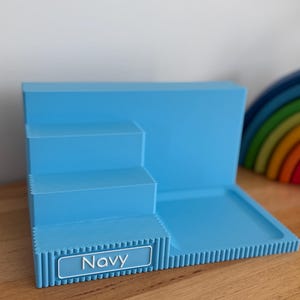 May include: A light blue tiered display stand with a flat rectangular base and three stepped levels. The front edge features a ribbed texture and a rectangular label with a white border containing the text Navy in white capital letters.