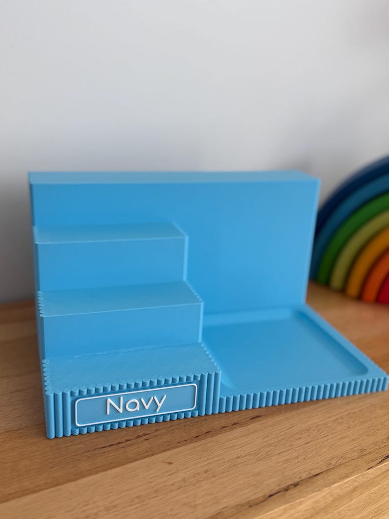 May include: A light blue tiered display stand with a flat rectangular base and three stepped levels. The front edge features a ribbed texture and a rectangular label with a white border containing the text Navy in white capital letters.