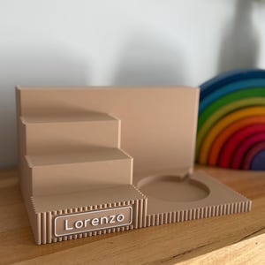 May include: A beige tiered display stand featuring a stepped design on the left and a circular cutout on the right. The front edge has a ribbed texture and a rectangular nameplate with the white text Lorenzo printed in a clean sans-serif font.