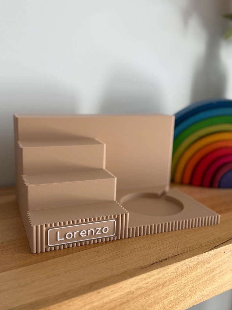 May include: A beige tiered display stand featuring a stepped design on the left and a circular cutout on the right. The front edge has a ribbed texture and a rectangular nameplate with the white text Lorenzo printed in a clean sans-serif font.