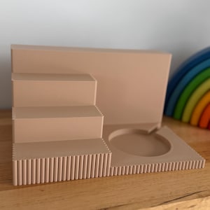 May include: A light beige display stand featuring a three-tiered staircase design with a fluted texture on the front edges. The base includes a circular cutout on the right side, and the entire structure is made of a solid, matte-finished material.