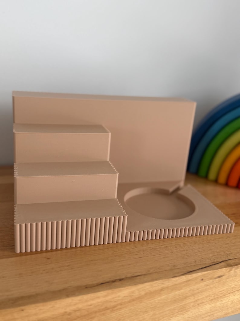 May include: A light beige display stand featuring a three-tiered staircase design with a fluted texture on the front edges. The base includes a circular cutout on the right side, and the entire structure is made of a solid, matte-finished material.