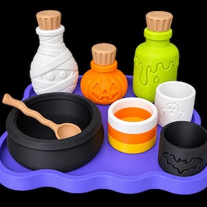 May include: A purple tray holding a black cauldron with a wooden spoon, a white mummy bottle, an orange pumpkin bottle, a green slime bottle, a candy corn cup, a white ghost cup, and a black bat cup, all featuring wooden-coloured bottle stoppers.