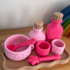 May include: A set of pink silicone play items including a bowl with a spoon, two bottles with tan stoppers, two small cups, and a star-topped wand, all arranged on a tan tray. The items feature embossed celestial and nature-themed motifs.