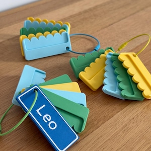 May include: A collection of rectangular plastic luggage tags in blue, green, and yellow, featuring scalloped edges and a smooth finish. One tag is prominently displayed with the name Leo printed in white text, attached to a green wire loop.
